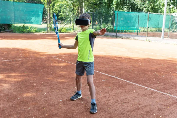 Online tennis training of boy in VR glasses standing on tennis court and preparing to make kick. New reality in sport, training in cyberspace.