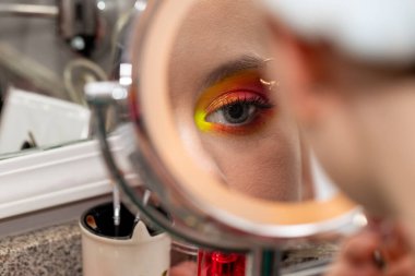 Genç bir kadın aynaya bakar. Gözündeki renkli makyaja bakar..