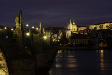 Vltava Nehri 'ndeki Charles Köprüsü ve üzerinde heykeller ve Çek Cumhuriyeti' nin merkezindeki Prag 'ın gece karanlığında sokak lambaları ve lambaların ışığı ve gökyüzünde bulutlar var.