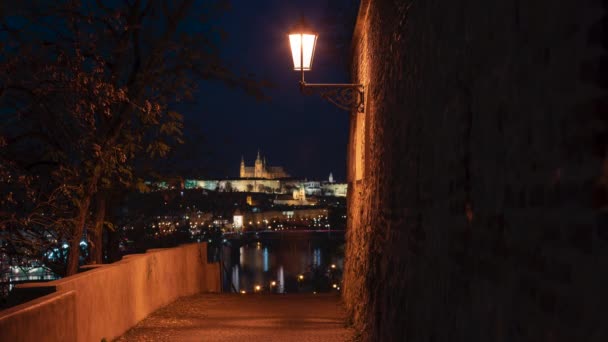 街路灯や歩行者用舗装された歩道からの時間経過光がプラハ城や聖ヴィート教会を照らし出します 夜にヴルタヴァ川と車のライトを流れる — ストック動画