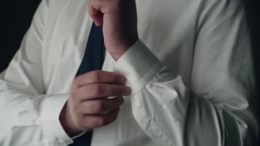 Beyaz gömlekli ve mavi kravatlı beyaz bir adam evin penceresinde duruyor ve sağ eli gömleğinin koluna bir düğme takıyor. Yakın plan.
