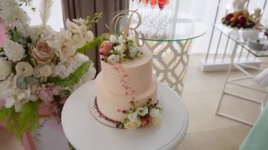İki katlı lezzetli düğün pastası pembe detaylarla süslenmiş, güller ve Latin harfi şeklinde bir süs büfenin arka planında duruyor, şampanya bardakları ve çiçek dekoru.