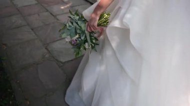 Şık, gösterişli bir elbisesi olan beyaz bir gelin ve uzun bir peçesi olan enerjik bir şekilde yolda yürüyor ve elinde farklı çiçek ve bitkilerden oluşan güzel bir düğün buketi tutuyor.