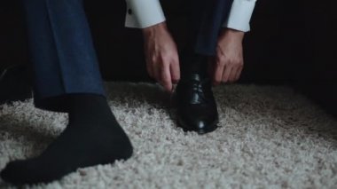 Beyaz gömlekli, mavi pantolonlu, siyah çoraplı beyaz bir adam pencerenin yanındaki bir koltukta ve büyük yün bir halıda oturuyor. Deri ayakkabı ve kravat bağcıkları takıyor.