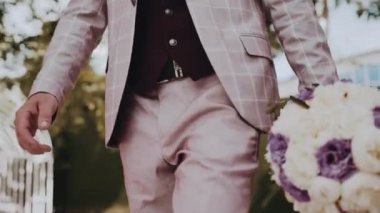 Beyaz kareli takım elbiseli, beyaz gömlekli, kahverengi yelekli, siyah kuşaklı beyaz bir adam dışarıda yürüyor ve elinde muhteşem bir buket taze beyaz ve mor şakayık taşıyor.