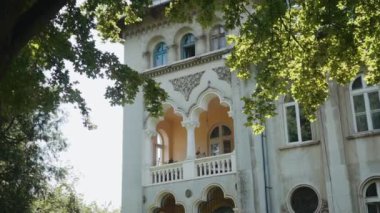 Geniş balkonu ve sütunları olan üç katlı, antika bir ev. Ağaçların dallarındaki yeşil yapraklardan, güneşli bir günde berrak bir gökyüzünün arkaplanından