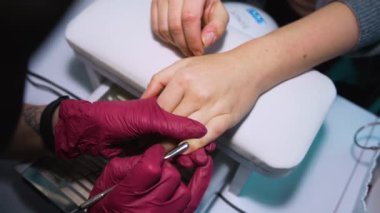 Azot eldivenli beyaz bir manikürcü kız sterilize edilmiş bir alet tutuyor - bir spatula ve bir güzellik salonundaki müşterinin sol elinin tırnağındaki ve yüzük parmağındaki tırnağı kaldırıyor.