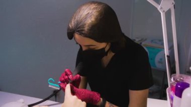 Üniformalı manikür ustası, tek kullanımlık maske, koruyucu başlık ve nitril eldivenler masasındaki güzellik salonunda oturuyor ve masa lambasının altında donanım manikürü yapıyor. Değirmen makası müşterinin tırnaklarını temizler