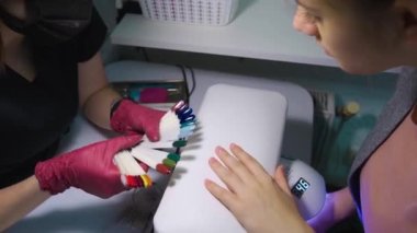 Manikür ustası müşteriye parlak vernikli bir palet gösterir ve müşteri sevdiği rengi seçer ve aynı zamanda elini ultraviyole lambada tutar.