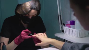 Manikür ustası bir güzellik salonunda tüm kurallara göre giyinir bir masada oturur ve bir masa lambasının altında müşterinin tırnaklarındaki jel cilasının altına bir taban uygular. Ortalama plan