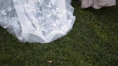Sarkık dantelli beyaz elbiseli gelin yeşil çimlerin üzerinde yürüyor ve yanında bej renkli ipek elbiseli kız arkadaşları var.