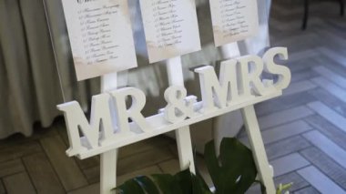 Lokantada dört beyaz tahta bacağın üzerinde duran cam kaplı bir standın içinde düğün ziyafetinde konuklar için oturma planı var ve aşağıda Latince mektuplar yazılmış Bay ve Bayan.