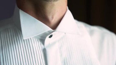 Kafkasyalı genç adam pencerenin yanında duruyor ve zarif bir şekilde siyah düğmeli beyaz bir gömleğin yakasını ayarlıyor.