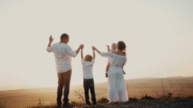 Gün batımında mutlu bir aile. Anne baba ve oğul ufukta el ele tutuşuyorlar ve ebeveynler çocuk büyütmekle meşguller. Kameraya el sallayan ve sonra babasına hava öpücüğü gönderen bir kadın.