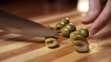 Aşçı kız, mutfaktaki tahta bir tahtada keskin bir mutfak bıçağıyla yeşil zeytinleri ikiye bölüyor. Bir yemek malzemesinin yakın plan çekimi.