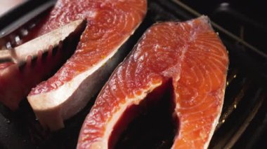 Erkek bir şef elektrikli ızgarada somon bifteği hazırlar ve metal mutfak maşasıyla balık parçalarını çevirir. Adam bifteğin çiğ tarafını yere koyuyor ve tepesinde çizgili sulu kahverengi kabuk var.