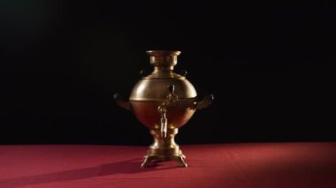 Masa örtüsünün üzerindeki siyah arkaplanda suyu ısıtmak için tasarlanmış ve Rus kültürüyle ilişkilendirilmiş eski metal semaver bulunuyor. Olağanüstü bir ev eşyası üzerinde kameraya vurmak