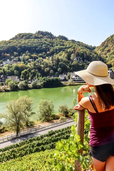 Young solo traveling woman with sun hat standing in a vineyard and looking over the hills and the Mosel river in Germany on a sunny day.