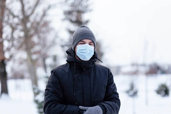 A young guy in winter wears a mask. Young man in protective antiviral mask on the street. The guy in winter clothes in a protective mask. Young man in warm clothes and scarf on a winter day.