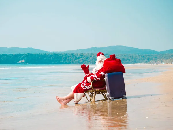 Santa Claus se sienta en una silla descalza con una maleta y una bolsa roja con regalos descansando en la orilla del mar —  Fotos de Stock