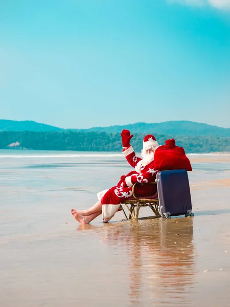 Santa con una maleta y una bolsa con regalos se sienta en la playa en un viaje — Foto de Stock