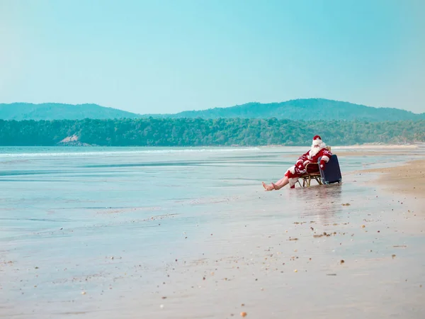 Feliz santa claus sentado en la orilla del mar con una maleta y con una bolsa roja bajo el sol descalzo levantando la mano —  Fotos de Stock