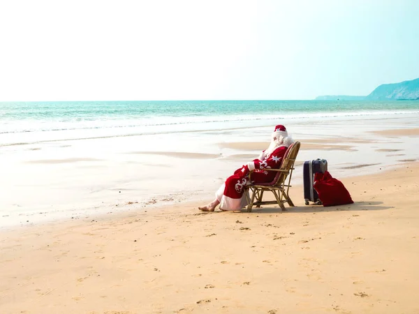 Święty Mikołaj siedzi na krześle boso z walizką i czerwoną torbą z prezentami spoczywającymi na brzegu morza. — Zdjęcie stockowe