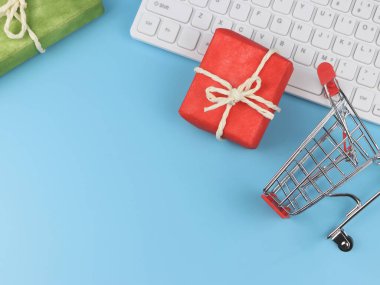 Kırmızı ve yeşil hediye kutularının üst görüntüsü, bilgisayar klavyesi ve mavi arka planda fotokopi alanı olan alışveriş arabası. İnternetten alışveriş ve Noel tatili konsepti.