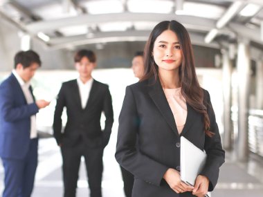 Resmi takım elbiseli Asyalı güzel iş kadını portresi elinde bilgisayar bilgisayarı, takımının önünde duruyor, gülümsüyor ve kameraya bakıyor..