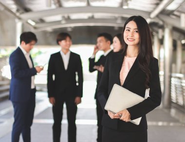 Resmi takım elbiseli Asyalı güzel iş kadını portresi elinde bilgisayar bilgisayarı, takımının önünde duruyor, gülümsüyor ve kameraya bakıyor..