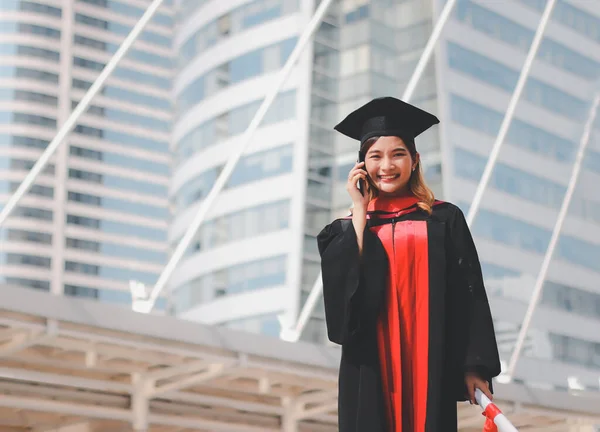 Happy Asian  graduated women in graduation gowns , holding diploma, smiling and talking on mobile phone. City building background.Education, successful concept.