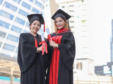 Mezuniyet elbisesi içinde kolları çaprazlanmış, gülümseyen ve kameraya bakan iki mutlu mezun kadın. Şehir binasının arka planı.