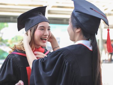 Mezuniyet elbisesi giymiş, birbirine yardım eden, mutlu bir şekilde gülümseyen iki neşeli mezun kadın. - Evet. Eğitim, başarı ve arkadaşlık kavramı.