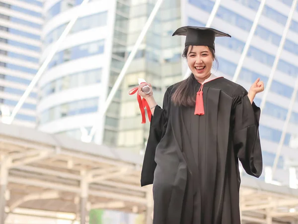 Happy graduated woman in graduation gowns holding diploma and raise fist, smiling happily. City building background. Education and successful concept.