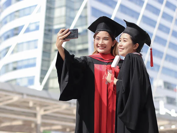 Two happy graduated women in graduation gowns making selfie photo with mobile phone. City building background. Education, Friendship and technology concept..