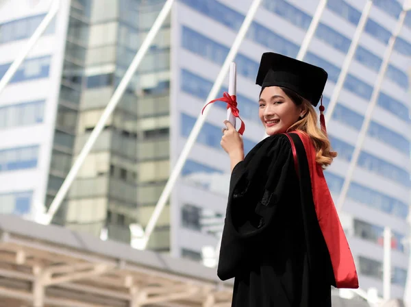 Happy Asian  graduated women in graduation gowns , holding her diploma, turning  back and smile at camera. City building background. Education and successful concept.
