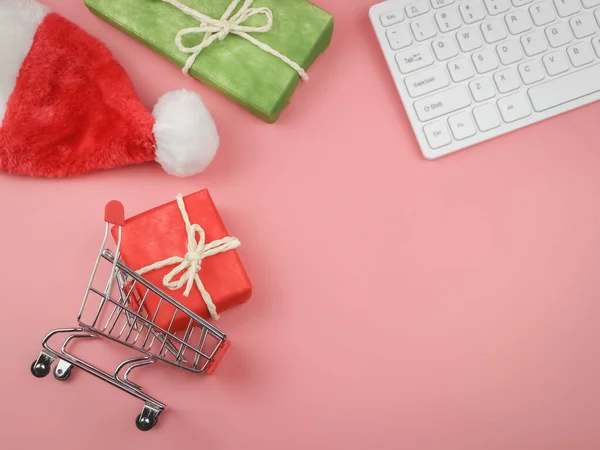 Alışveriş arabasındaki kırmızı hediye kutusu, yeşil hediye kutusu, Noel Baba 'nın şapkası ve pembe arka plandaki bilgisayar klavyesi. Noel tatili online alışveriş konsepti.