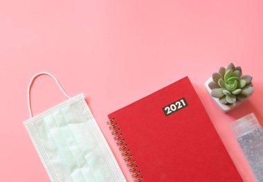Top view of red diary or planner 2021 with medical face mask, alcohol sanitizer hand gel and succulent plant pot on pink background. New year 2021 planning, COVID19 prevention and new normal concept. clipart