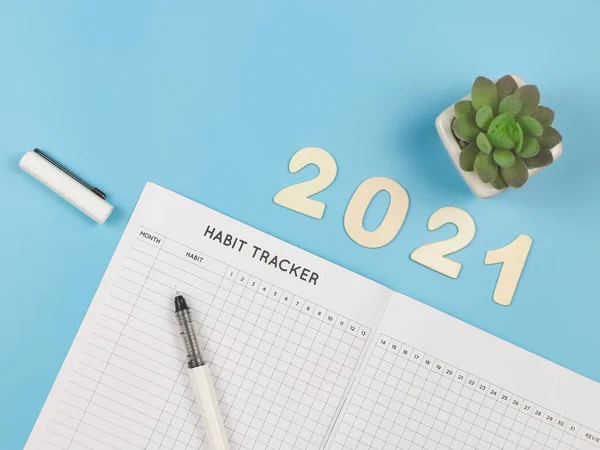 Top view or flat lay of habit tracker book with pen, wooden number 2021 and succulent plant pot on blue background.