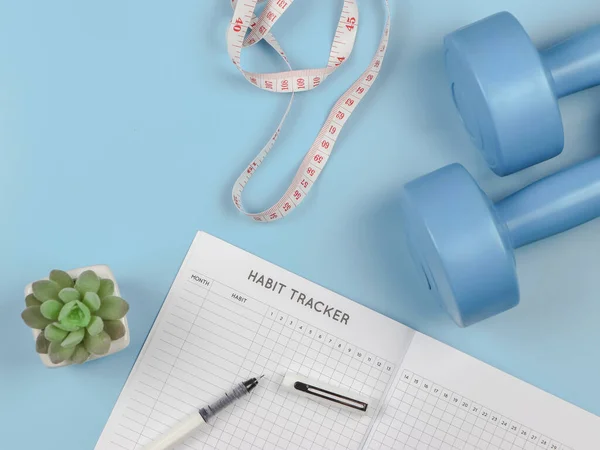 Top view or flat lay of habit tracker book, dumbbells, measure tape and succulent plant pot on blue background with copy space. lose weight, exercise, self development.