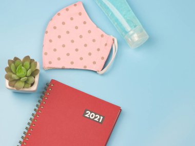 Kırmızı günlük veya planlamacı 2021 'in üst görüntüsü pembe benekli kumaş maskesi, alkol el jeli ve mavi arka planda bitki saksısı. Yeni yıl 2021 planlama, COVID19 önleme ve yeni normal kavram.
