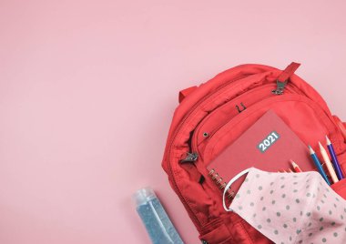 Okul gereçleri, kumaş maskesi ve alkol dezenfektanı ile dolu sırt çantasındaki kırmızı 2021 günlüğünün üst görüntüsü. Kopya alanlı pembe arka planda. COVID-19 önleme, 2021 'de okula geri dönüş ve yeni normal.