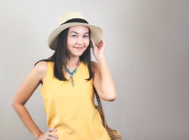 Sarı kolsuz gömlek giyen Asyalı bir kadının portresi, örülmüş çanta taşıyan bir şapka, beyaz arka plan, gülümseyen ve kameraya bakan. Yaz konsepti.