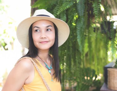 Asyalı bir kadının sarı kolsuz gömlek giydiği, şapkasının örülmüş çantasını tuttuğu, bahçede dikildiği, yana bakan görüntüsü. Yaz konsepti.