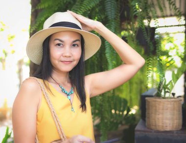 Asyalı bir kadının sarı kolsuz gömlek ve şapka giydiği, örgü çantalı, bahçede dikilip kameraya baktığı görüntüyü kapatın. Yaz konsepti.