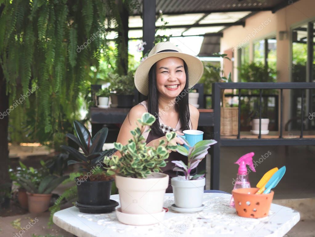 Portrait of happy Asian woman wearing hat and sleeveless shirt sitting in the garden holding blue cup of coffee,  smiling happily and looking at camera.