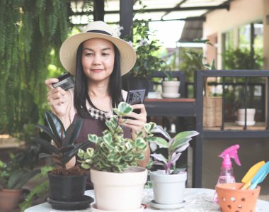 Şapka ve önlük takan Asyalı bahçıvanın portresi, kredi kartıyla alışveriş yapan bitkiler. Bahçede oturan Asyalı kadın elinde cep telefonu ve kredi kartıyla