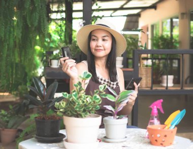 Şapka ve önlük takan Asyalı bahçıvanın portresi, kredi kartıyla alışveriş yapan bitkiler. Bahçede oturan Asyalı kadın elinde cep telefonu ve kredi kartıyla