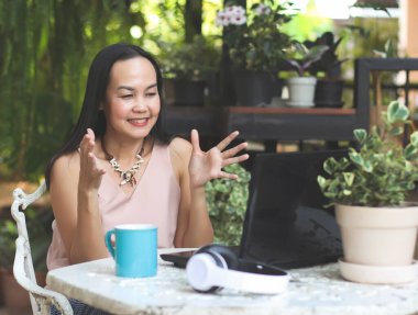 Asyalı kadın portresi evdeki bahçesinde oturuyor, bilgisayar defteriyle video görüşmesi yapıyor, bilgisayar ekranında konuşuyor ve işaret veriyor..
