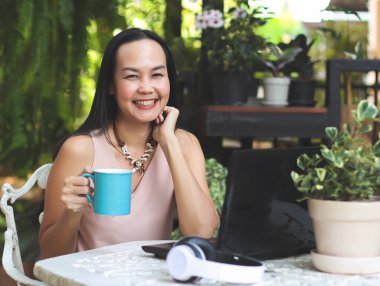 Asyalı bir kadının portresi evinin bahçesindeki masada oturuyor, elinde mavi kahve fincanı, gülümsüyor ve kameraya bakıyor. Bahçede kahve içen kadın..